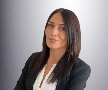 Woman with straight dark hair wearing a black blazer and white shirt, standing against a gradient gray background, looking at the camera with a neutral expression.
