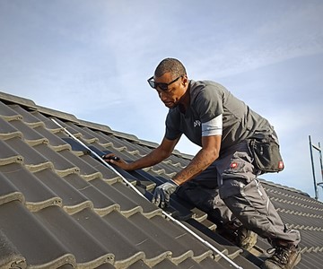 A worker wearing safety glasses measures roof tiles while kneeling on a sloped roof under a clear sky.