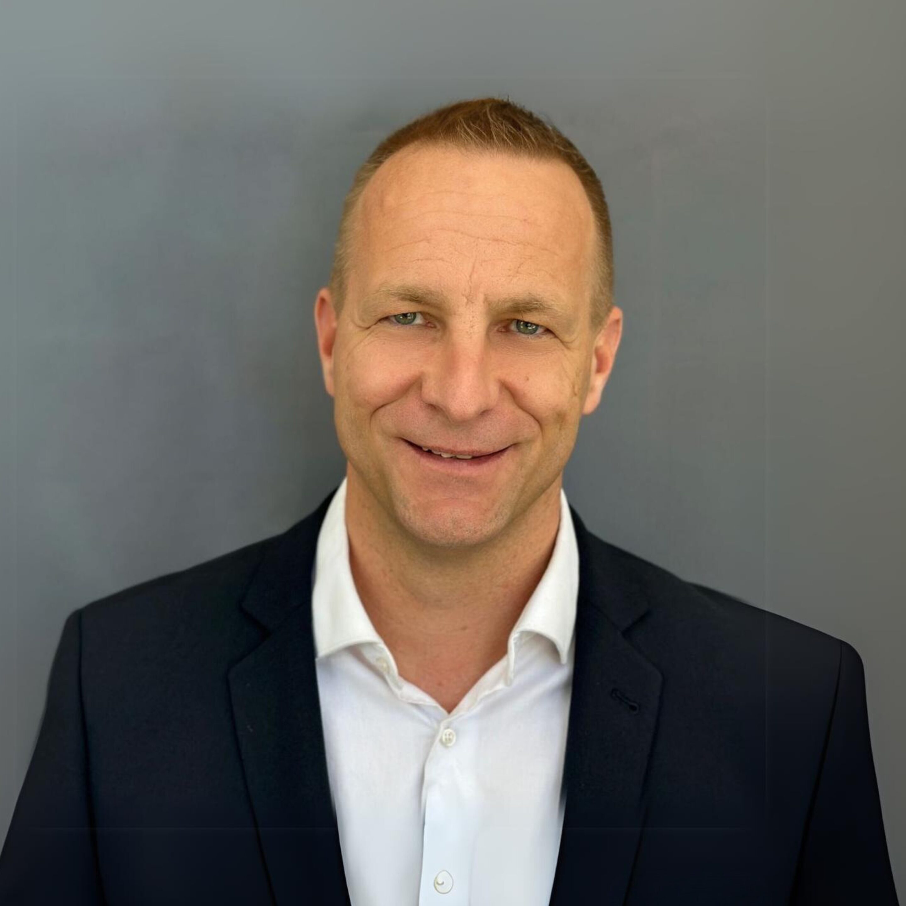 A man wearing a dark suit jacket and white shirt stands in front of a plain gray background, looking at the camera and smiling.