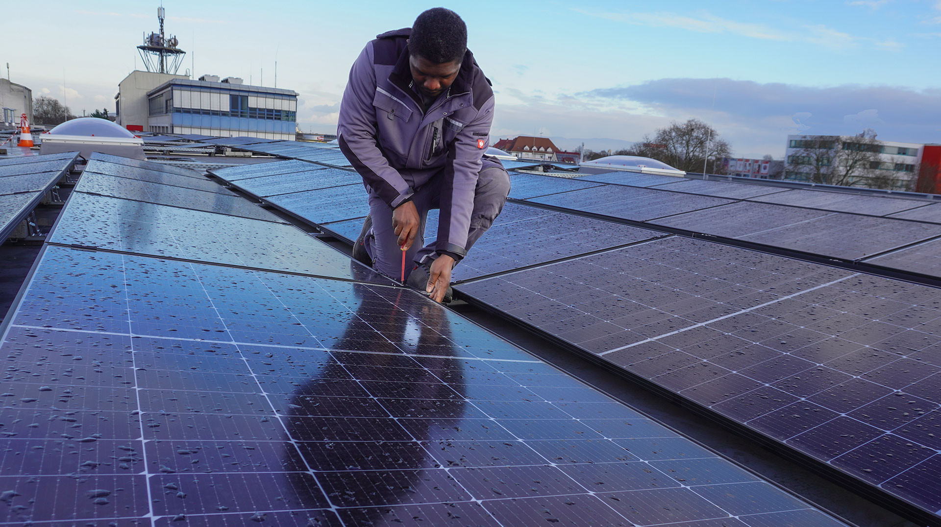 Eine Person in Arbeitskleidung justiert oder überprüft Solarmodule auf einem Dach. Auf den Modulen sind Wassertropfen zu sehen, im Hintergrund sind Gebäude zu sehen.